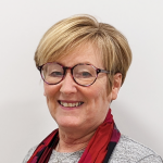 White lady with short blonde hair and glasses wearing a grey top with a red scarf.