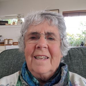 Older white lady with short grey hair and glasses sitting on a sofa wearing a flowery top.