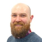 White man with close shaven head and full ginger beard and moustache, with hazel eyes. Wearing bright checked shirt under a blue jumper.