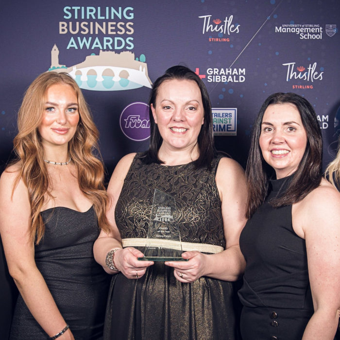 Picture of 5 white females in formal wear holding the award for Charity of the Year in front of a backdrop advertising the Stirling Business Awards.