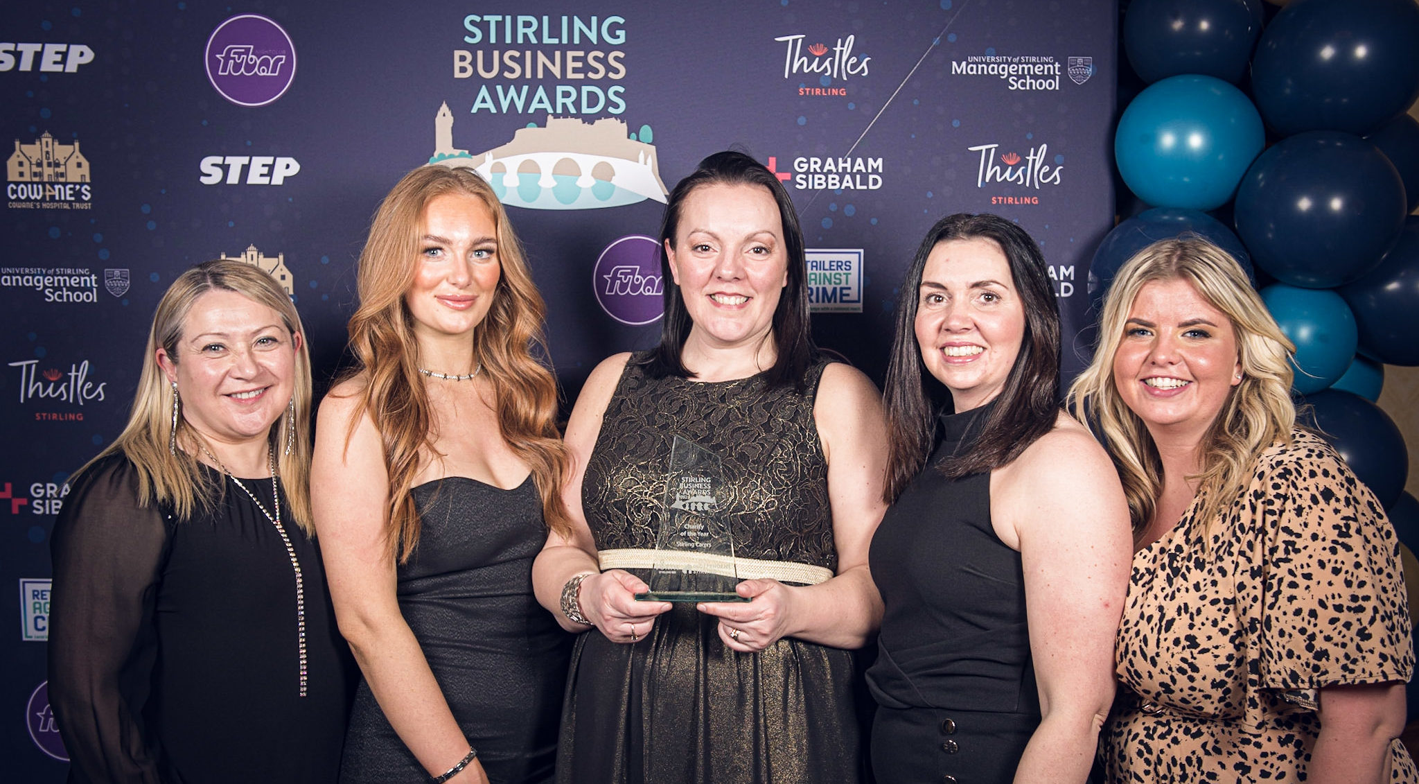 Picture of 5 white females in formal wear holding the award for Charity of the Year in front of a backdrop advertising the Stirling Business Awards.