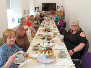 A long table with afternoon tea set out and surrounded by ladies enjoying the food.