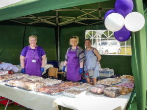 Three members of staff in a green gazebo preparing to serve food, standing behind trays of sandwiches.