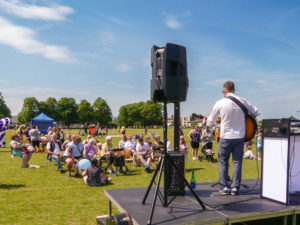 Singer with a guitar stood on a stage with a group of people sat enjoying the music in the sunshine.