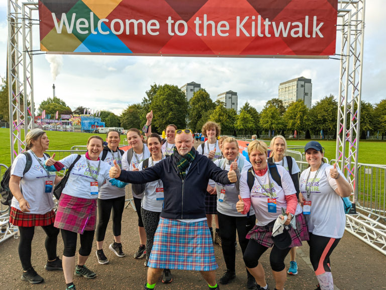 Group of people standing with Sir Tom Hunter, wearing Stirling Carers Centre t-shirts under the start line banner for The Kiltwalk.