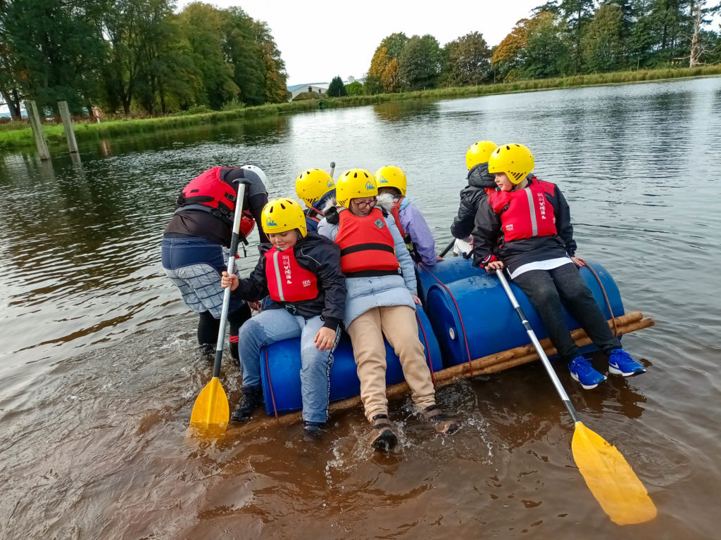 Young Carers wearing yellow helmets and red buoyancy aids floating on a river on a raft made of oil drums and bamboo canes.